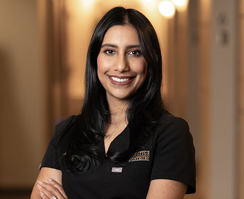 The image shows a woman standing confidently with her arms crossed. She has dark hair, is wearing a black shirt, and is smiling at the camera. Behind her, there s a blurred background that suggests an indoor setting, possibly a hallway or corridor.