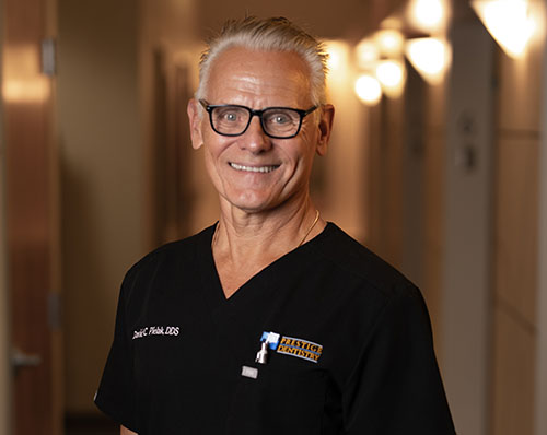 The image features a man standing in a hallway, smiling at the camera. He is wearing glasses, has gray hair, and is dressed in a white shirt with a black tie and a name tag that includes text.