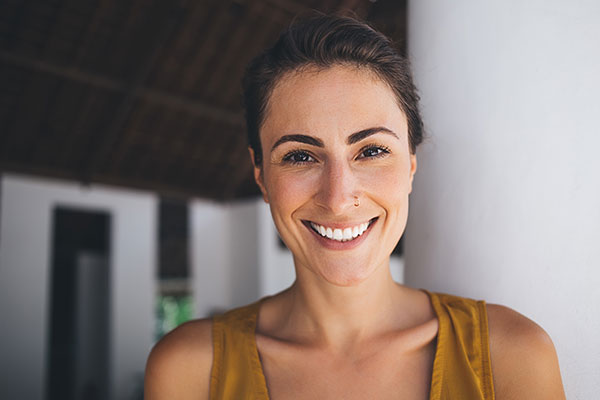 A young woman with a radiant smile stands against a white wall under a roof with dark beams, wearing a yellow top.