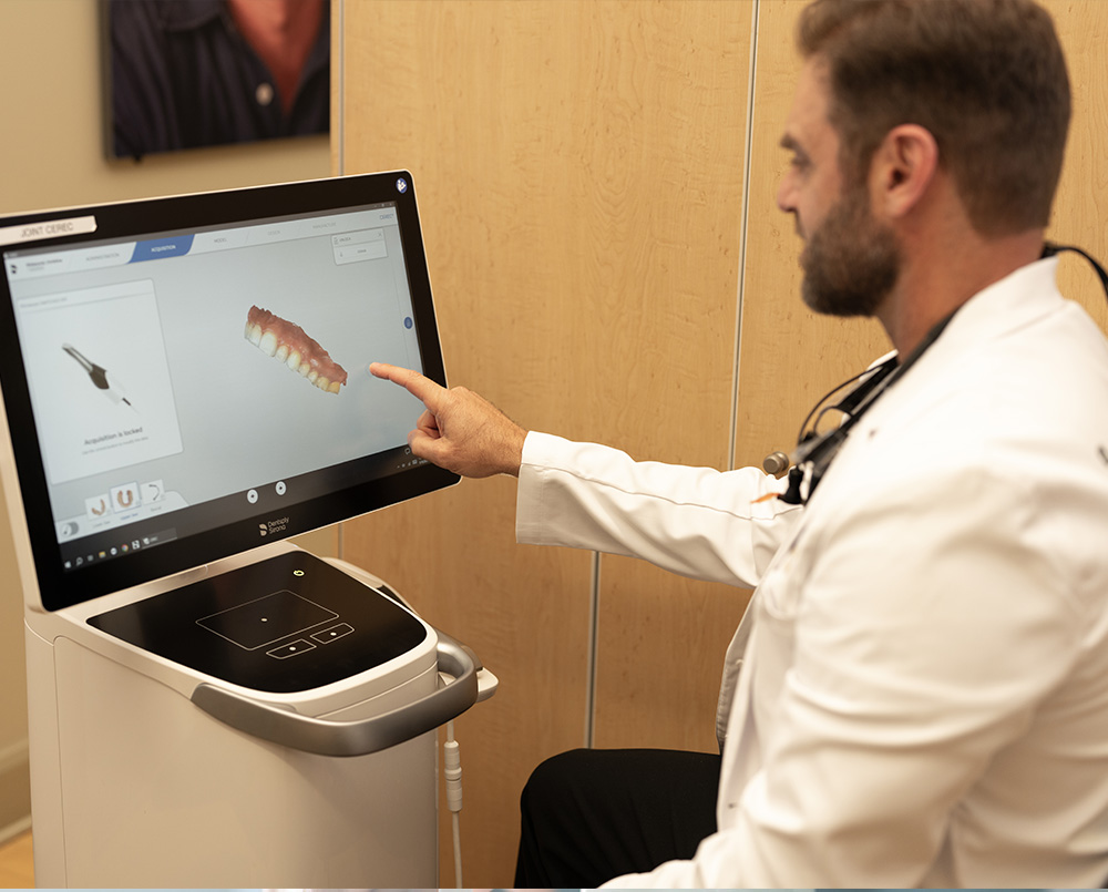 The image shows a man in a white coat standing at a computer screen displaying a 3D model of a human head, likely in a medical or educational setting. He appears to be examining or presenting the model.