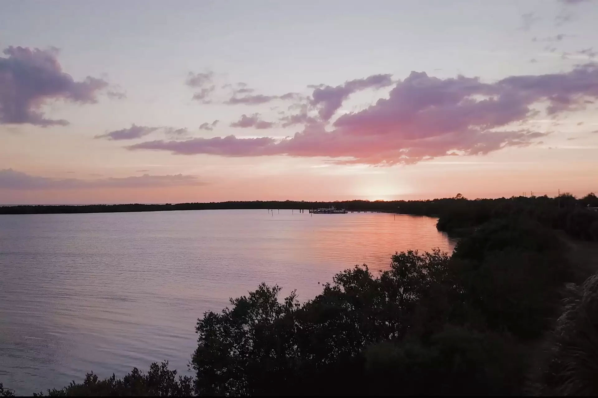 The image shows a serene sunset over a tranquil body of water with a shoreline visible on the right, featuring a clear sky with soft pastel colors reflecting off the water s surface.