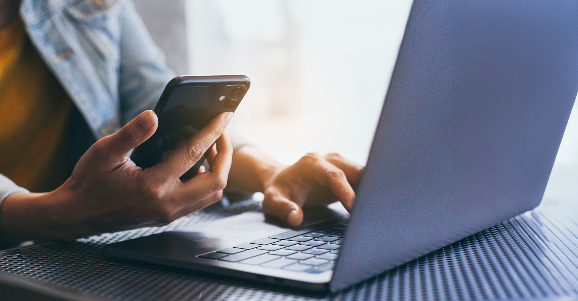 The image shows a person s hands typing on a laptop keyboard while they are sitting at a table with a smartphone in front of them, both devices are in focus against a blurred background that suggests an indoor setting.