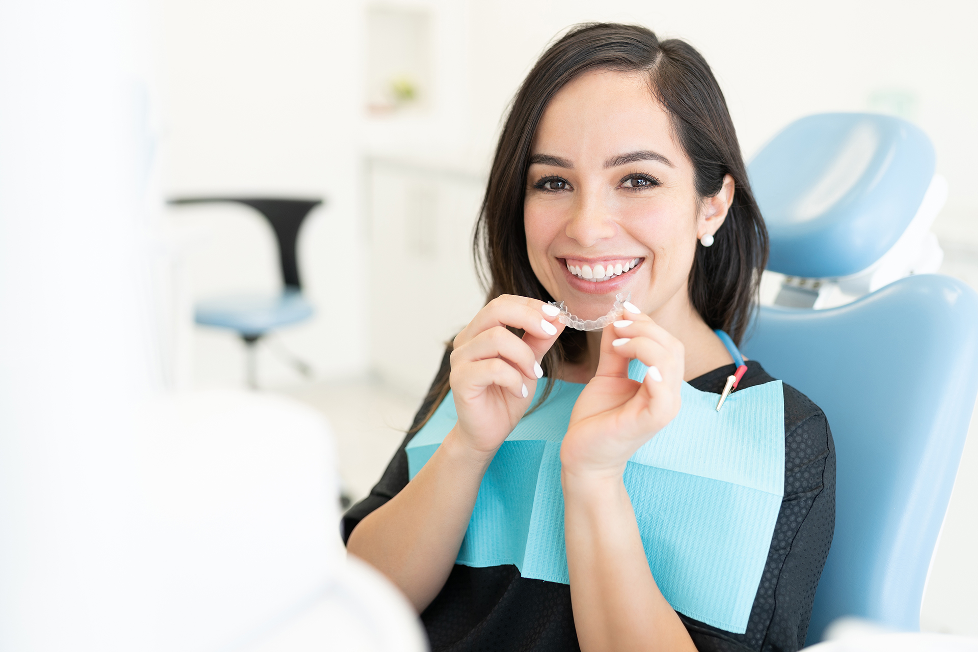 A woman sitting at a dental chair, smiling, holding a toothbrush, with dental equipment in the background.