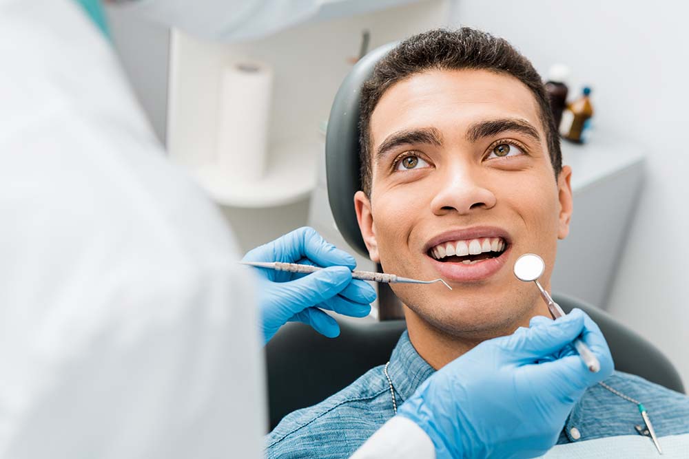 The image shows a person sitting in a dental chair with a smile on their face while receiving dental care, with a dental professional performing the procedure and wearing gloves and a surgical mask, set against a white background with dental equipment visible.