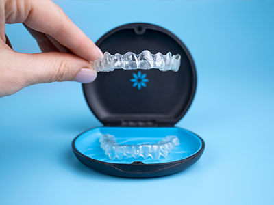 The image shows a person s hand holding an open box containing a set of clear braces with blue accents, placed on a blue background.