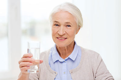 The image features an older woman holding a clear glass with water, smiling slightly, and raising the glass towards her mouth as if she s about to drink from it.
