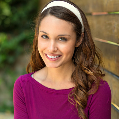 A young woman with long brown hair smiling at the camera, wearing a purple top and a headband with a bow.
