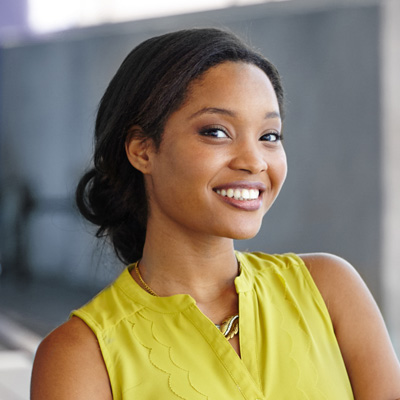 The image shows a smiling woman with dark hair wearing a yellow top and standing against a backdrop that includes a partial view of a building.