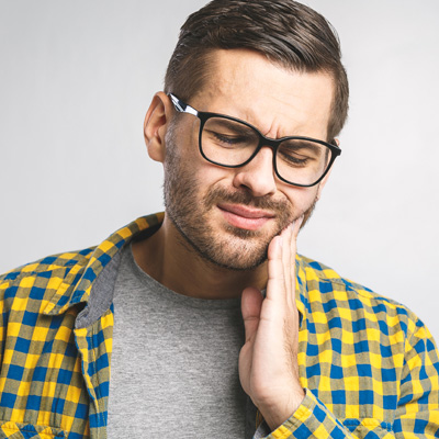 The image shows a man with glasses, wearing a yellow plaid shirt, holding his hand to his mouth as if he s in pain or discomfort.