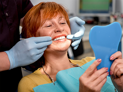 A woman with red hair sitting in a dental chair, smiling broadly while looking at her reflection in a mirror, with a blue dental model placed on her teeth.