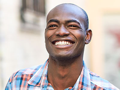 The image features a smiling man with short hair, wearing a plaid shirt and standing outdoors against a backdrop of a building with windows.