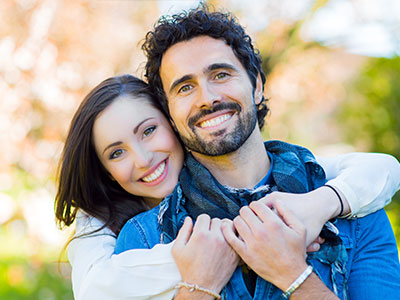 A man and woman are embracing each other outdoors during daylight  they both have beards and are smiling, with the man wearing a scarf around his neck.