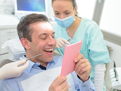A man sitting in a dental chair, holding a pink card with a surprised expression, while being attended to by a female dental professional.