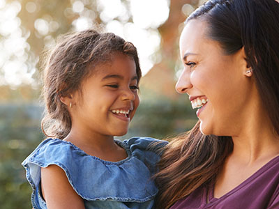 The image depicts a woman and a young child outdoors during daylight, with the woman smiling and looking at the child who is also smiling and appears to be laughing.