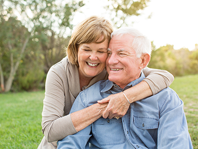 The image shows an older couple embracing each other outdoors during daylight, with both individuals smiling and appearing content.
