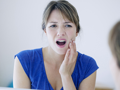 The image shows a woman with her mouth open, looking at her reflection in a mirror while brushing her teeth. She appears concerned or surprised, as indicated by her facial expression.