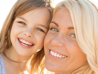 A woman and a young girl are smiling at the camera, with the woman s arm around the child, against a blurred background.