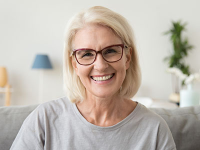 The image shows a woman with short blonde hair wearing glasses and a light-colored top, smiling at the camera.