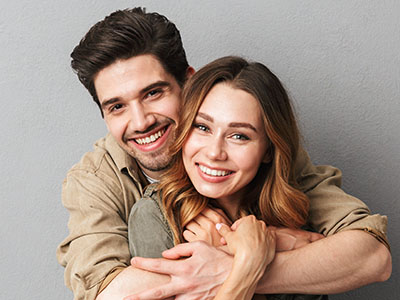 A man and woman are embracing each other in a close pose  the man has dark hair and a beard, wearing a light-colored shirt, while the woman has long hair, a neutral expression, and is dressed in a light-colored top with a V-neckline.