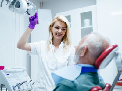 A woman in a white coat stands next to an older man with a medical device strapped to his head, both in a clean, sterile room with medical equipment and a purple glove on her hand.