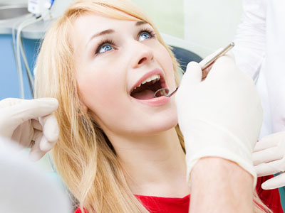 The image shows a woman sitting in a dental chair with her mouth open, receiving dental treatment, with a dentist wearing gloves and working on her teeth.