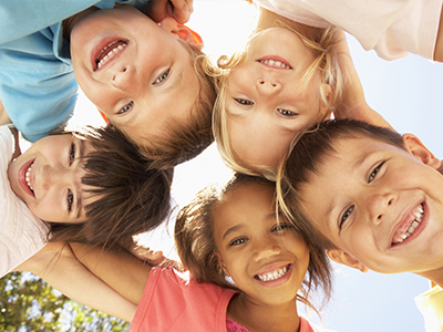 The image shows a group of children, possibly siblings, posing closely together with smiles on their faces, set against a bright outdoor background.