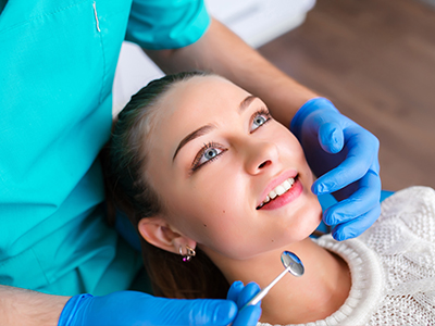 The image shows a person receiving dental care with a hygienist performing an examination using a mirror and a dental tool, while the patient smiles at the camera.