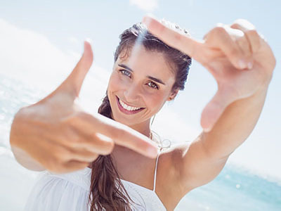 The image shows a smiling woman taking a selfie with her fingers framing the camera lens against a bright, sunny background.